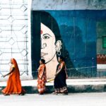 Three Indian women in traditional saris walk past a vibrant mural on a city sidewalk.